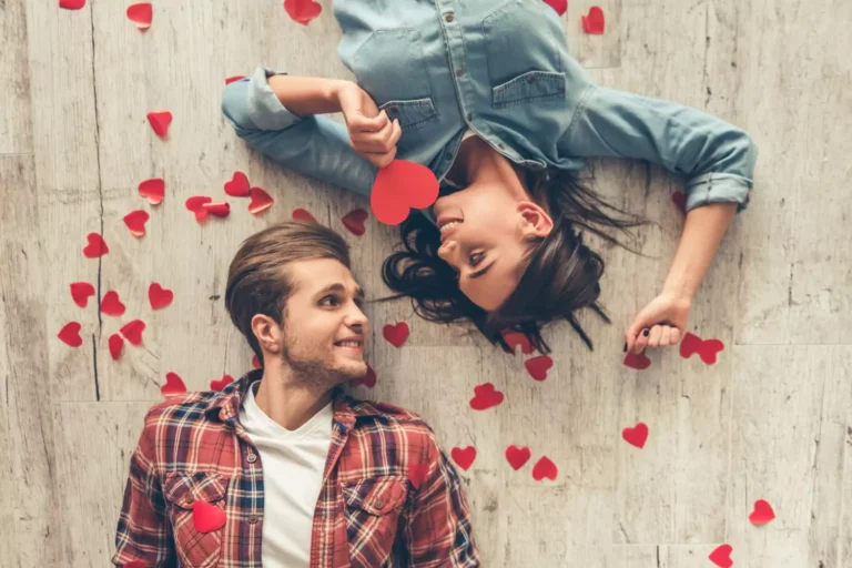 A couple lying on the floor surrounded by heart confetti, symbolizing the power of commitment in marriage, as they gaze into each other’s eyes with love and happiness.