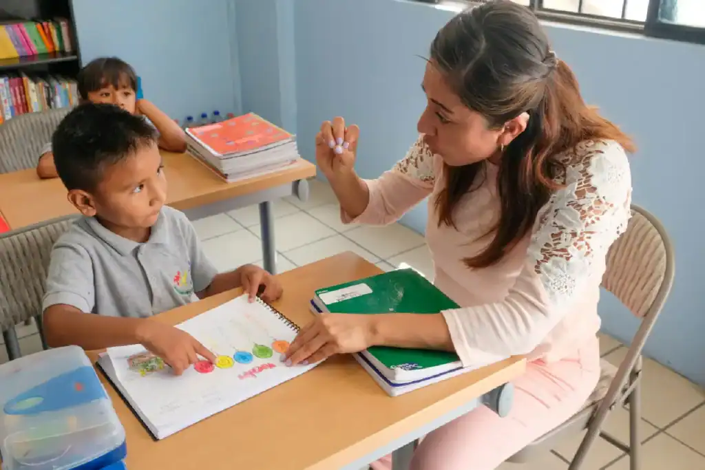 Teacher gestures while guiding a young boy with special education needs through a colorful workbook activity at a classroom desk.