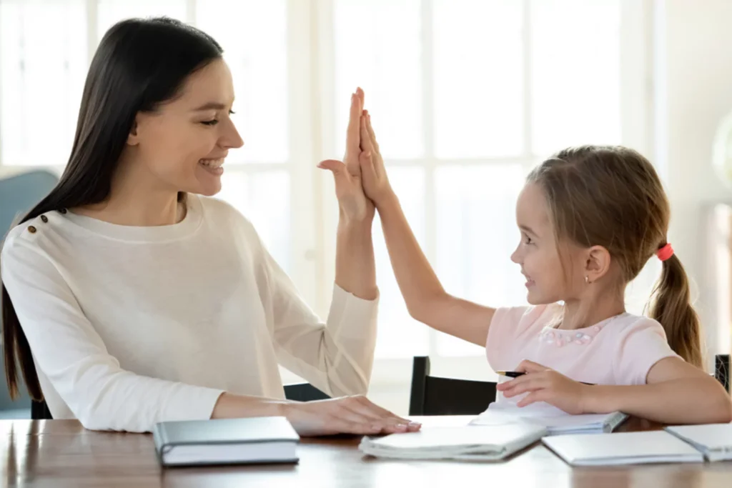A mother and daughter high-fiving each other while sitting at a table with books and notebooks, demonstrating effective home school strategies.