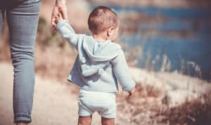 Shown from behind, a toddler boy holding mom's hand as they walk along the sandy shore of a lake