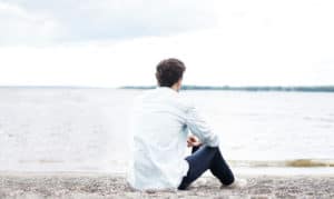 A man wearing jeans sits by himself near the beach.