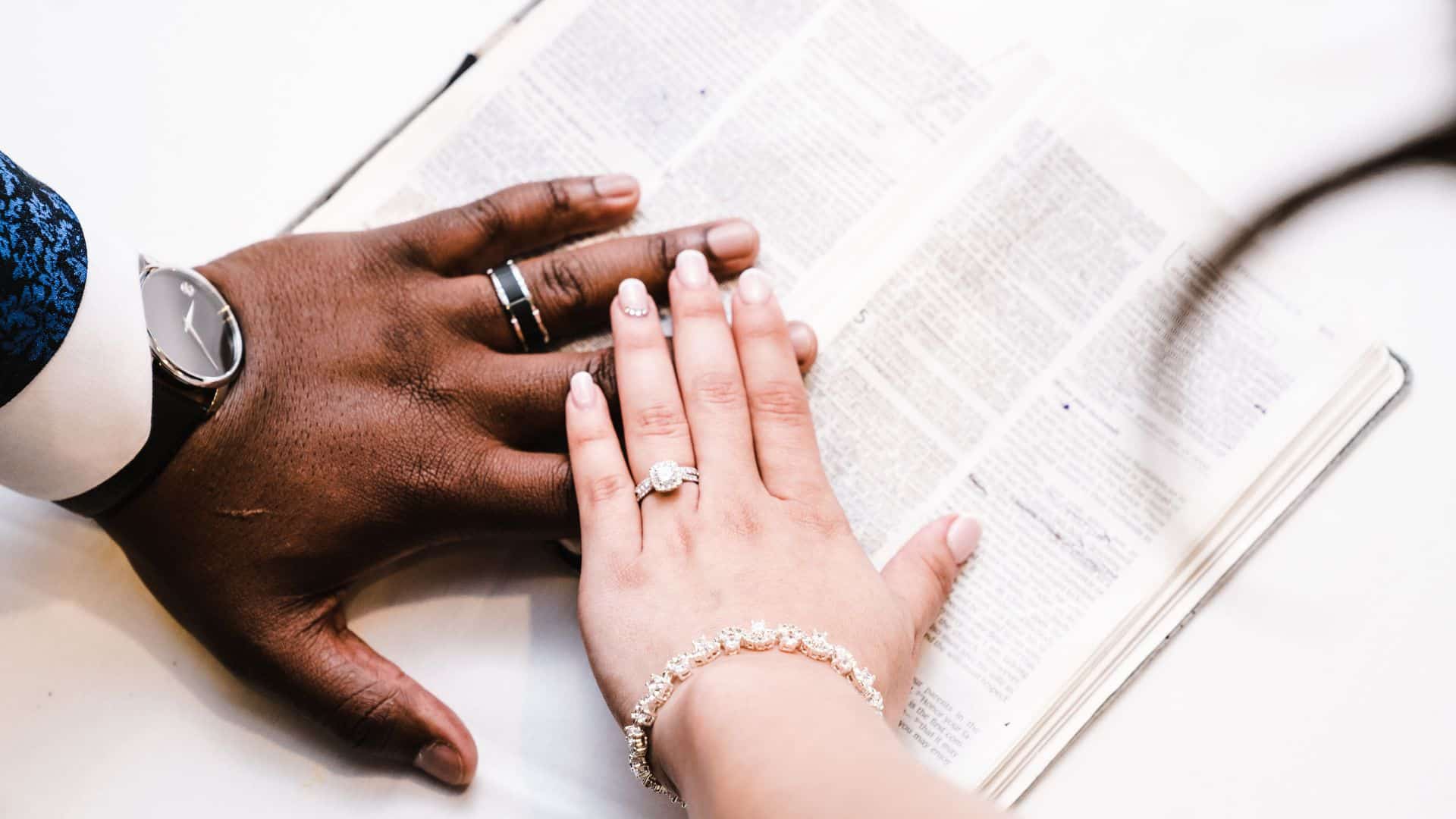 God's design for marriage. Two hands, a man's and a woman's, overlap as they touch an open Bible. They are both wearing wedding rings.