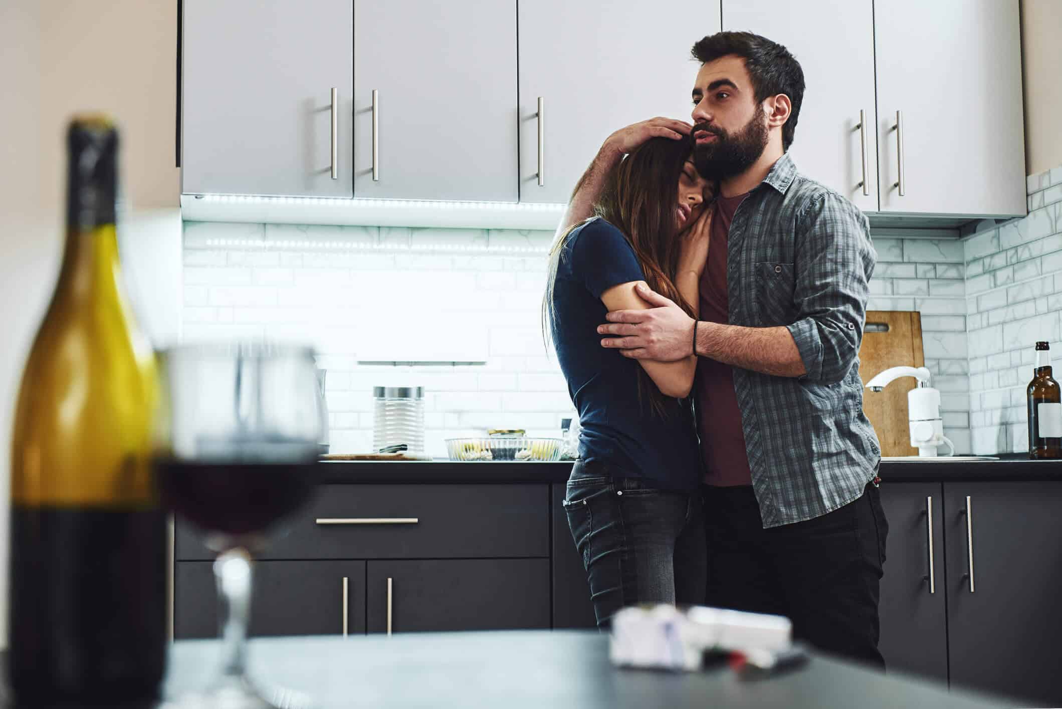 Concerned husband embracing his sad wife in their kitchen, with her head resting on his shoulder