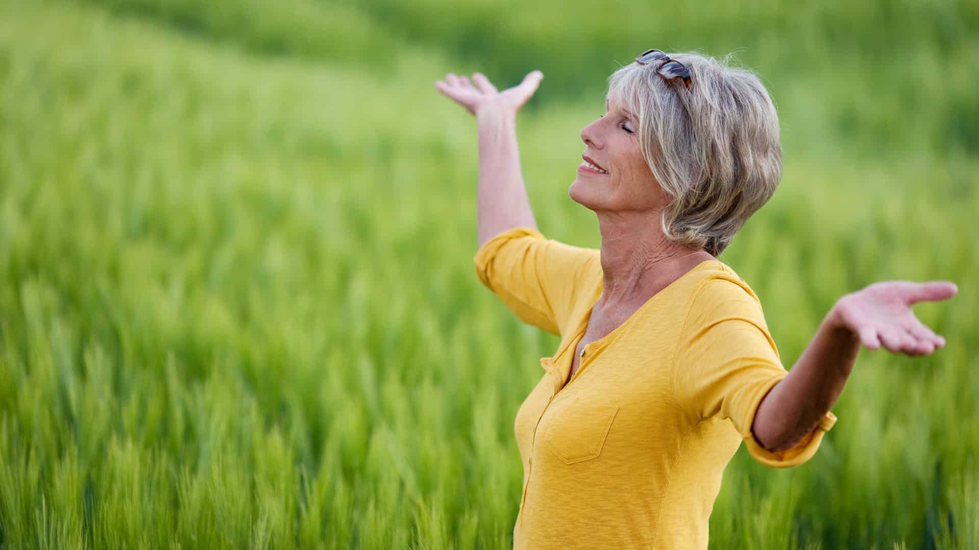 Smiling middle-age woman with outstretched arms standing in a pretty green field, enjoying nature