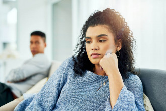 Young black woman in serious thought sitting on a couch, with a blurry background of a concerned man staring at her