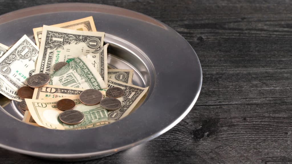 church collection plate with paper money and coins inside resting on dark wood table