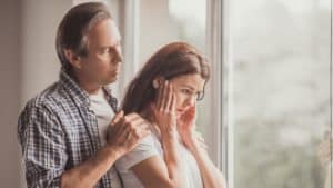 Middle-age husband and wife. She looks upset, he's standing behind her with his hands on her shoulders to comfort her.