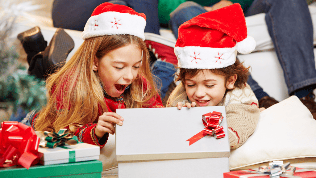 Two little girls in red Santa hats opening a present. They are surprised and grateful.
