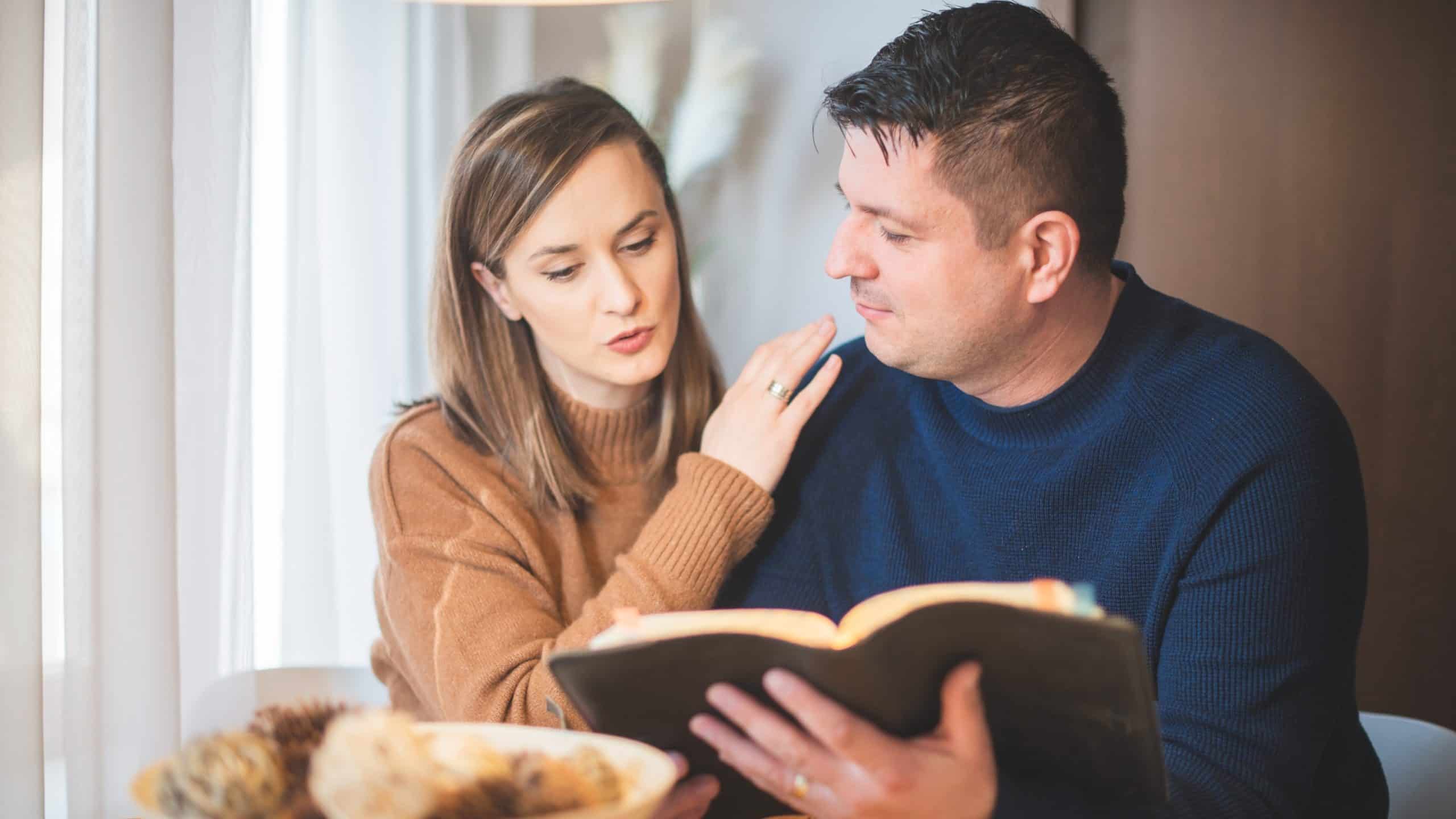 husband and wife sitting at table husband reading bible wife reading over his shoulder