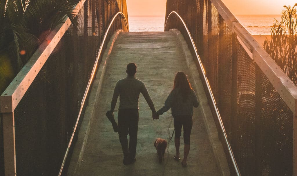 A couple walks on a bridge at sunset with their dog.