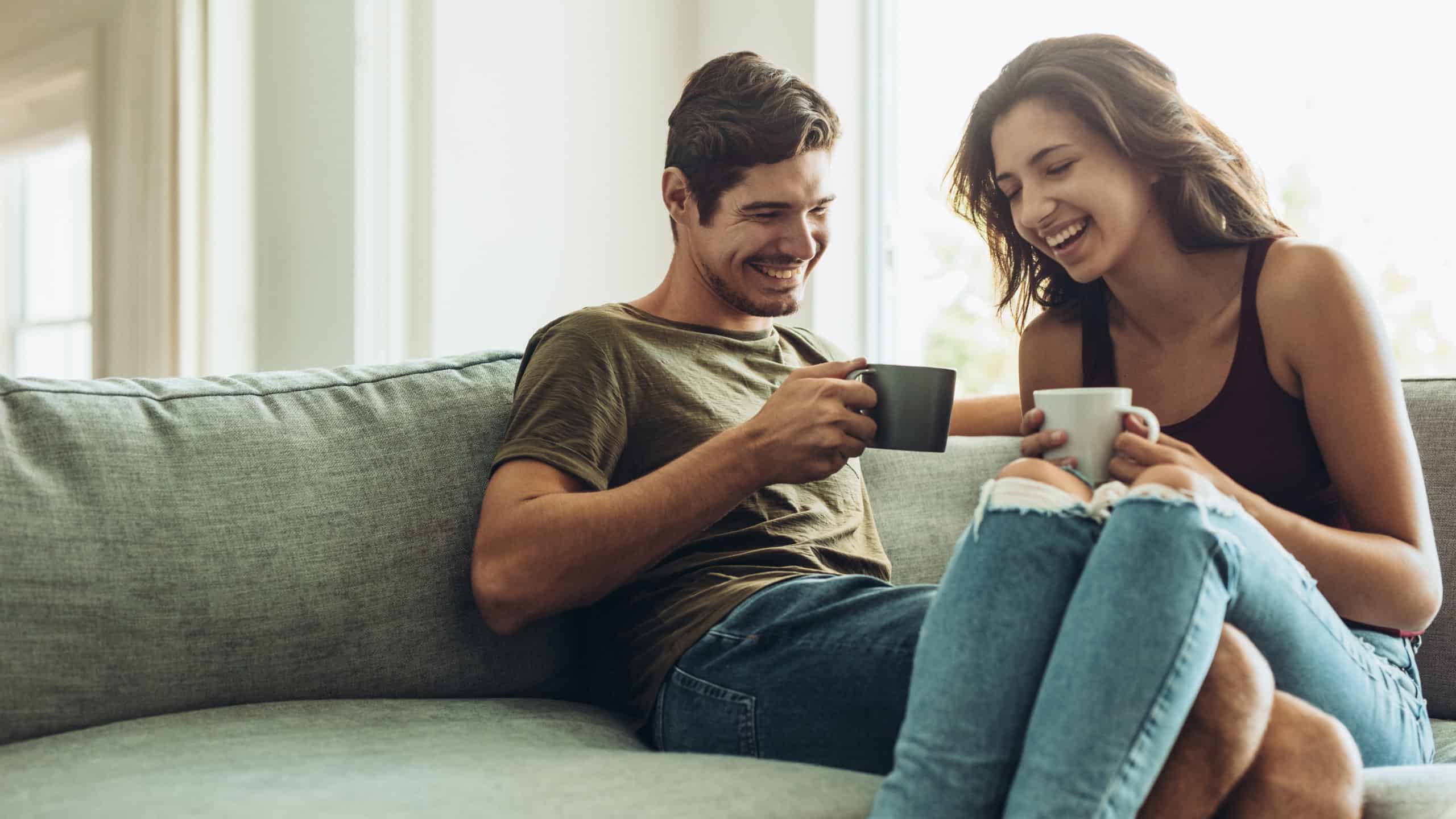 Couple sitting together on couch, laughing and enjoying cups of coffee
