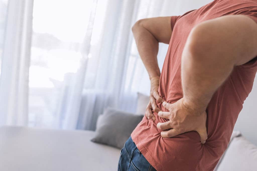 A man standing in his bedroom, gripping his lower back in pain