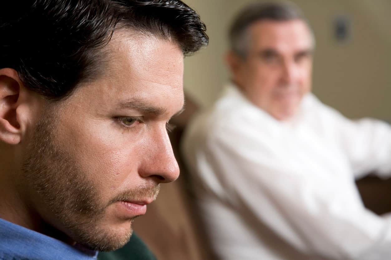 Close up profile of a serious looking, contemplative man, with his father sitting in the background looking at him