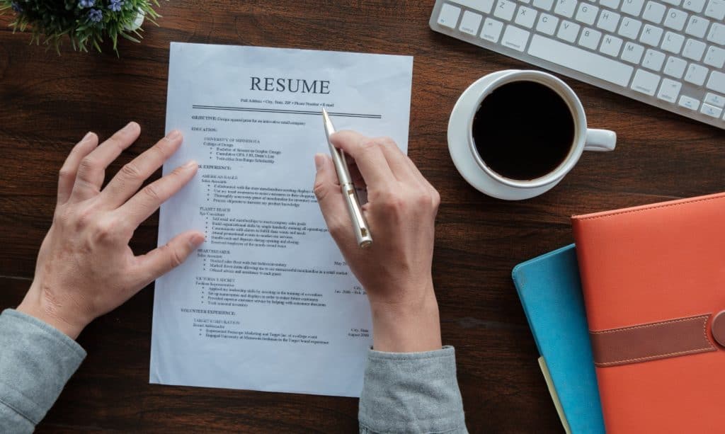 A man's hands, with one holding a pen and checking his resume while seated at a desk with a cup of coffee, shown from above