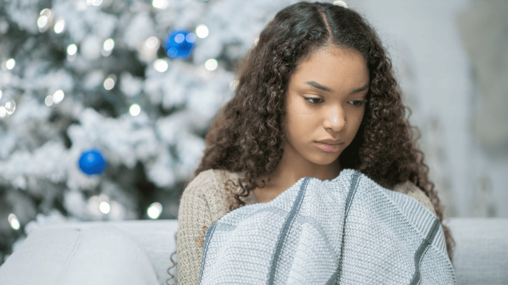 Handling the holidays after loss, a sad young woman sitting on a white coach holding a blanket, with a Christmas tree in the background.