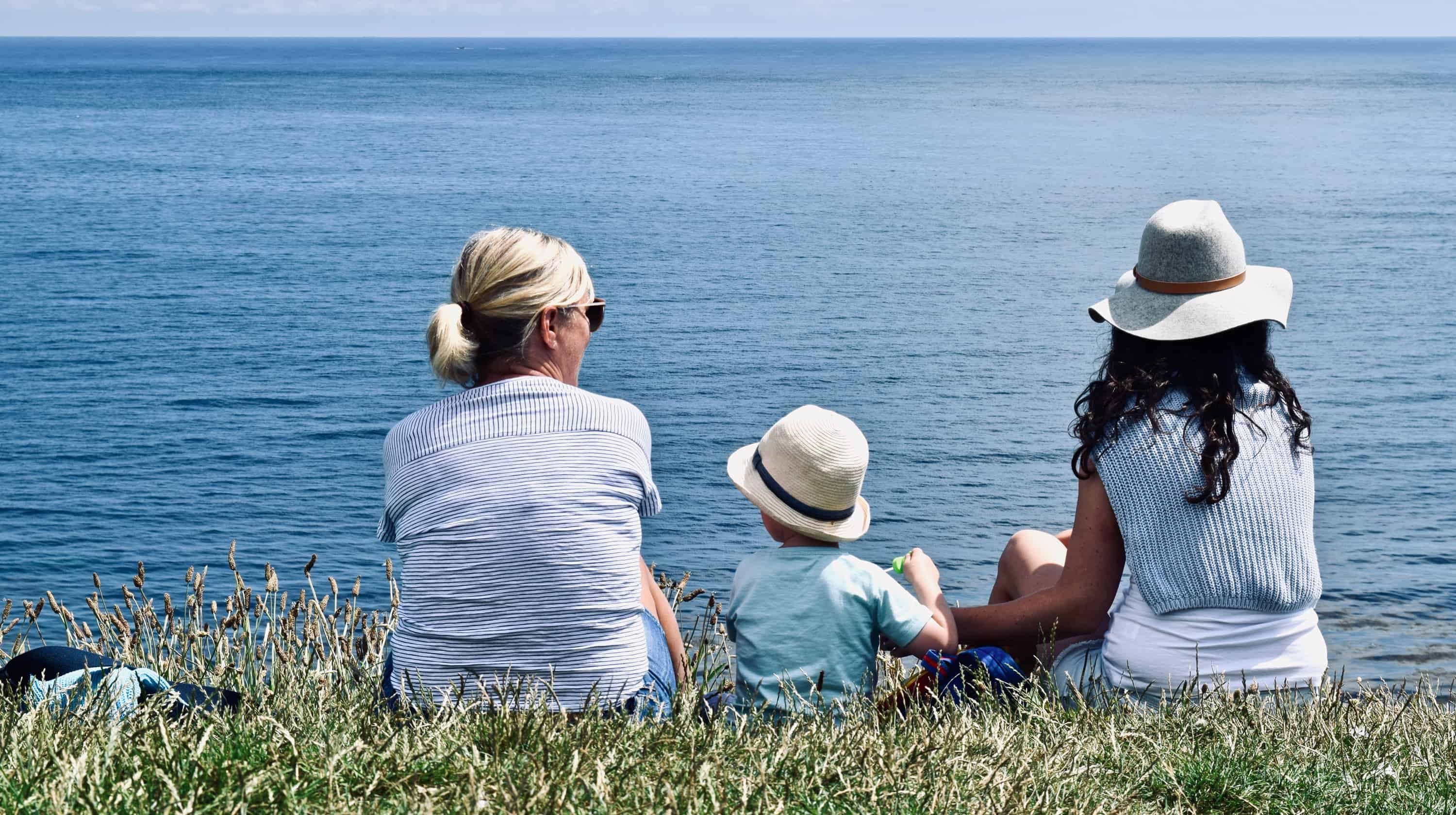 A grandmother, young child and young woman sit on a grassy hill overlooking the ocean.