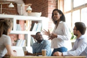 Young, confident black woman standing at a meeting table, making a presentation to her millennial co-workers