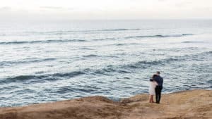 A couple walsk along a rocky shore near a beach.