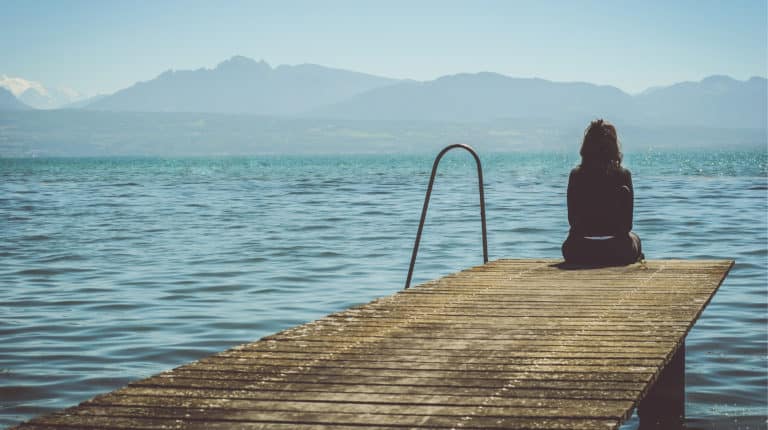 Shown from behind, a lone woman sitting at the edge of a dock overlooking a lake with mountains on the horizon