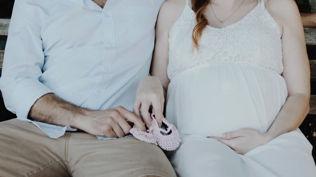 A pregnant woman sits next to her husband; together they're holding a pair of pink baby shoes.