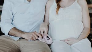 A pregnant woman sits next to her husband; together they're holding a pair of pink baby shoes.