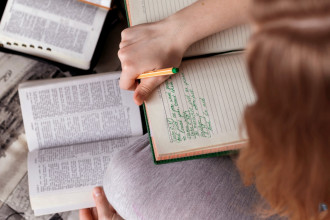 Shown from above, a woman sitting cross-legged taking notes as she reads a Bible