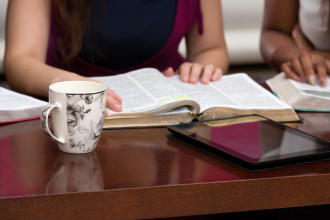 Close up of several open Bibles on a table with women's hands resting on them, indicating a Bible study