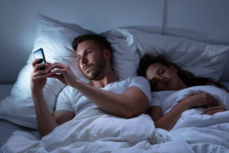 Man looking at his phone in bed while his wife sleeps next to him