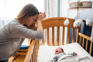 Young, stressed-out mom leaning against a crib with her newborn lying there crying