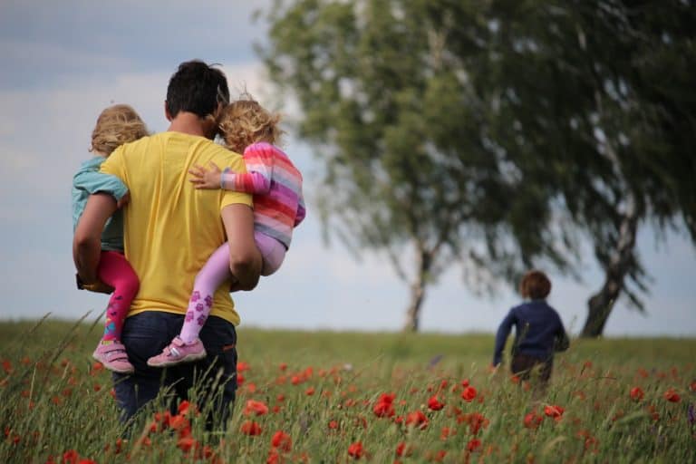 Dad holding his two daughters while walking through field of flowers; young son running in the background