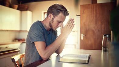 A man seated at a kitchen table praying before an open Bible