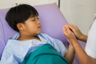 Close-up of sleeping, sick child in hospital bed, his hand being held by a nurse