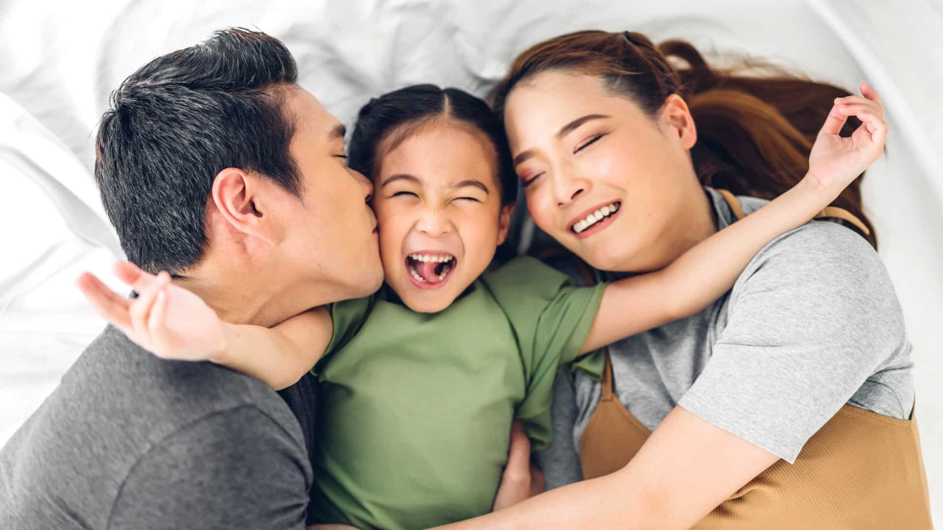 Joyful mom and dad hugging their laughing young daughter as they lie together on a bed