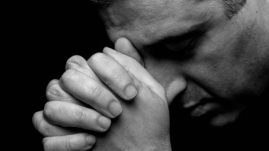 Black and white close up of man praying with eyes closed and head resting on his folded hands