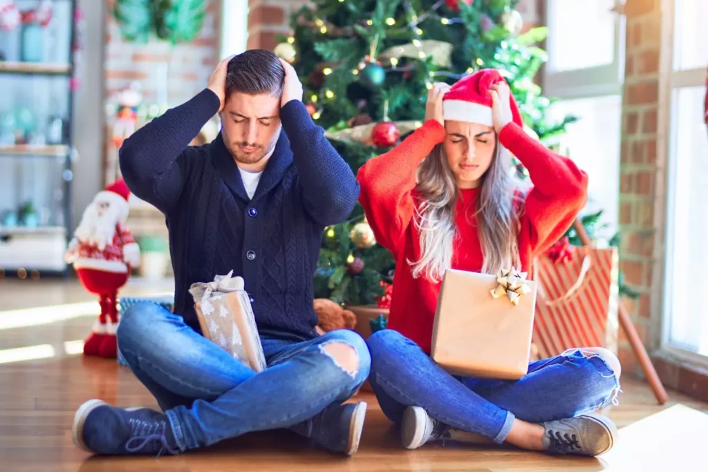 A married couple sits on the floor with their hands pressed against their heads, symbolizing the holiday stress that they are facing.