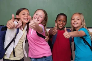 Group of four smiling kids with backpacks standing in front of a chalkboard. All are giving a thumbs-up to the camera.