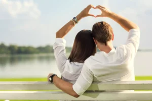 A couple sitting on a bench by the water, forming a heart shape with their hands, symbolizing the impact of marriage help in rekindling love and connection.