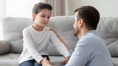 Dad kneeling down, having serious, compassionate talk with young son sitting on the couch