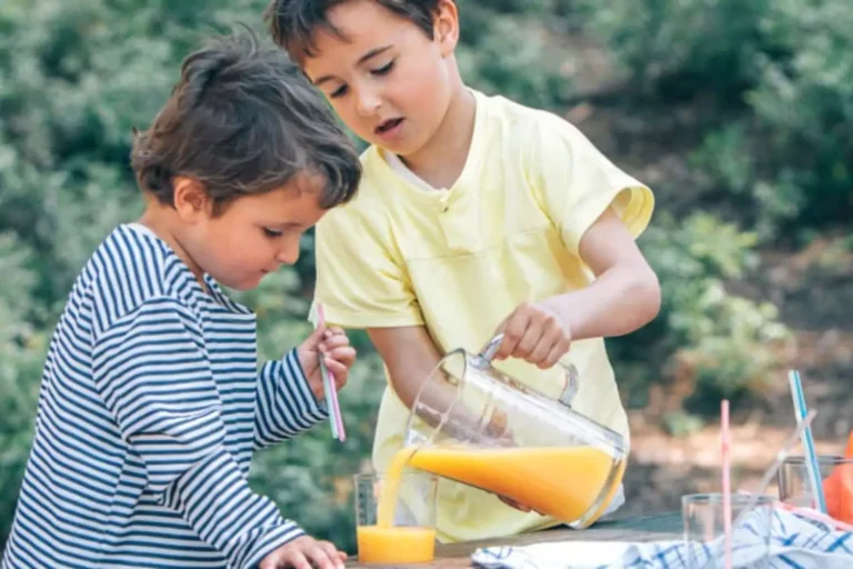 A young boy pouring a glass of orange juice for another boy at an outside table