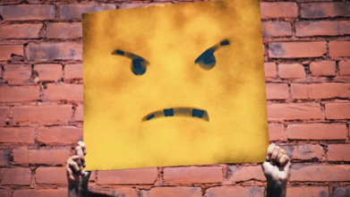 A grungy-looking yellow sign being held up by someone’s hands; sign depicts a close-up of an angry-faced emoji