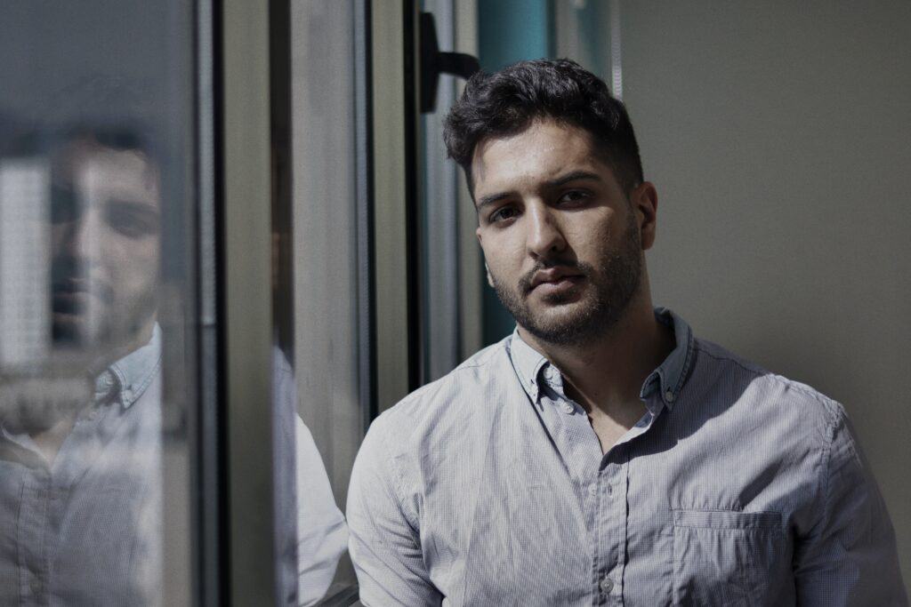 A man is standing by a window looking seriously at the camera