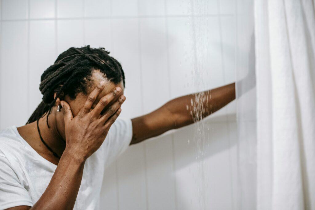 man standing in shower with clothes on hand over face regret