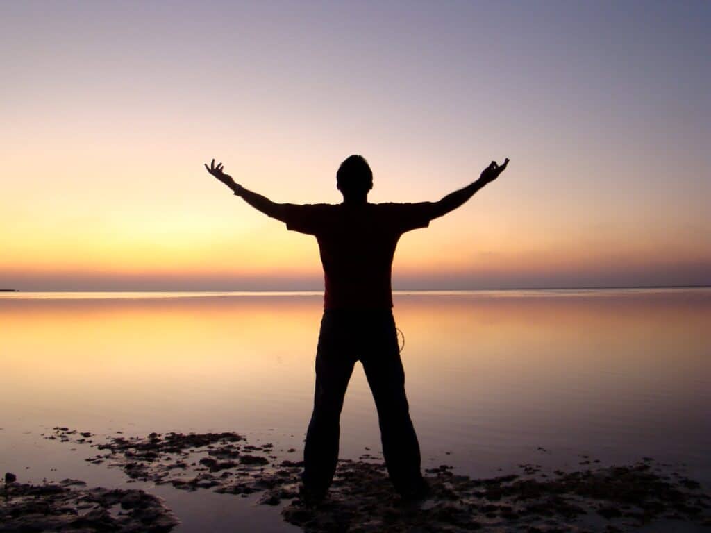 man holding up his arms at sunset over water