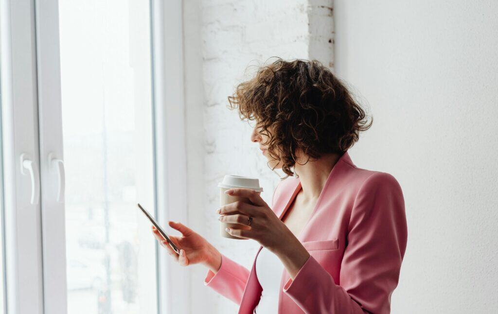 woman holding coffee looking at phone by window