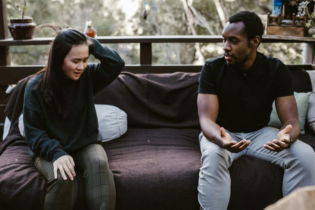 couple sitting apart on couch, man questioning woman sad