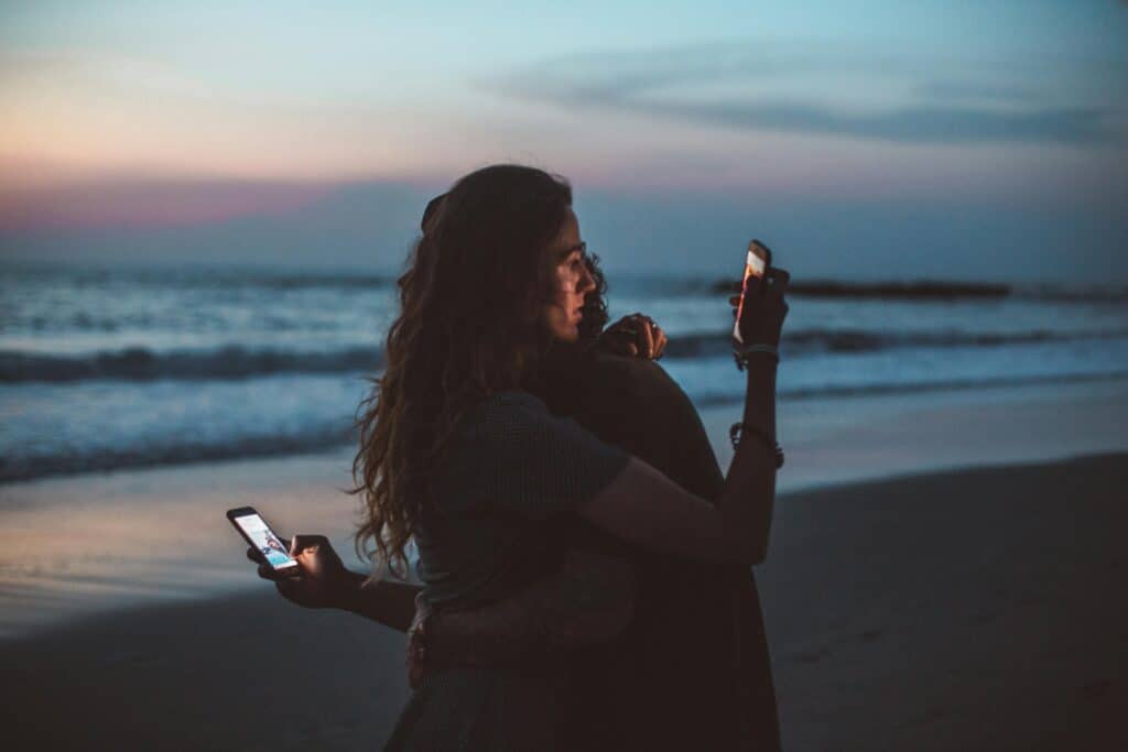couple hugging on beach both looking at phones