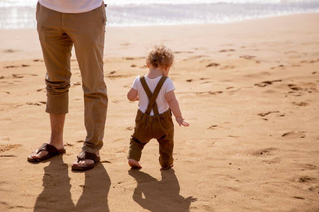 adult male legs toddler boy walking on beach