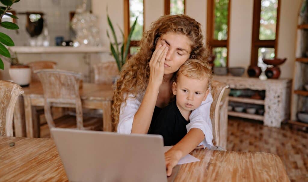 mom looking tired or sad sitting at table with computer toddler boy on lap