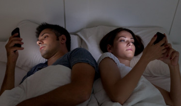 Husband and wife lying in bed, each staring intently at their phone
