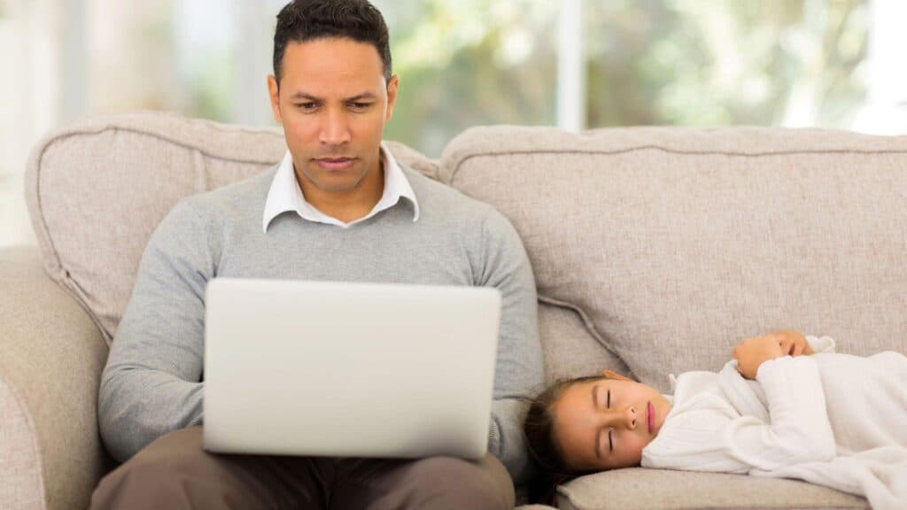 Man trying to create a work-life balance by working at home. Sitting on the couch with his computer in his lap and his daughter sleeping next to him